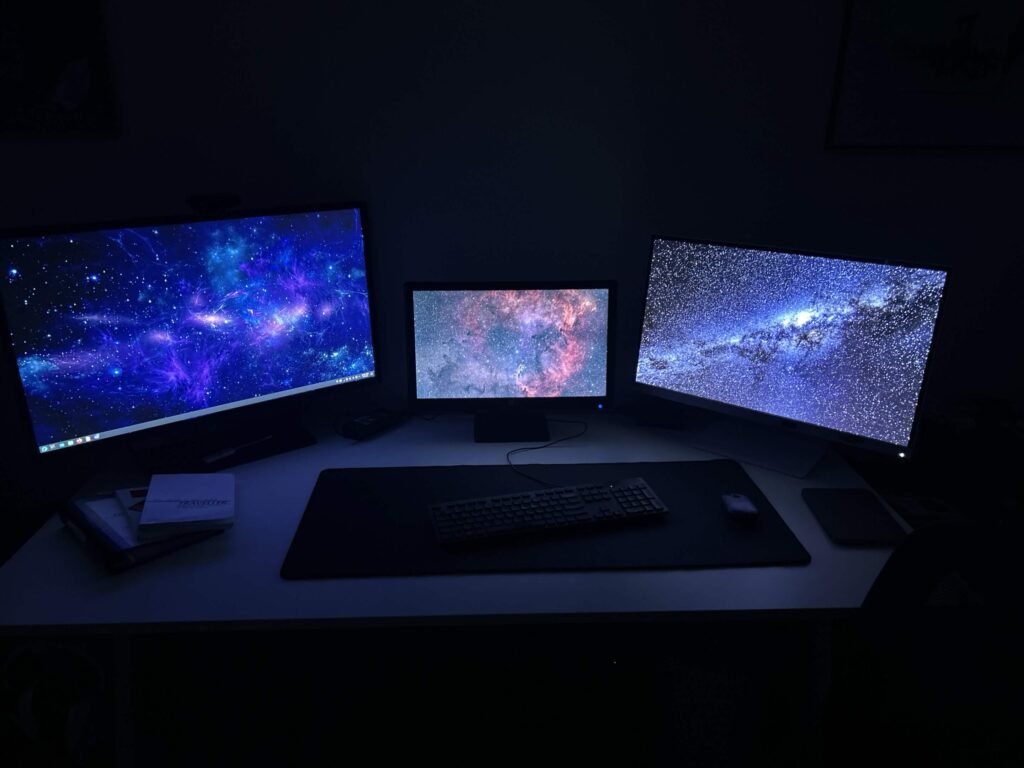 3 computer monitors on a desk, in a darkened room